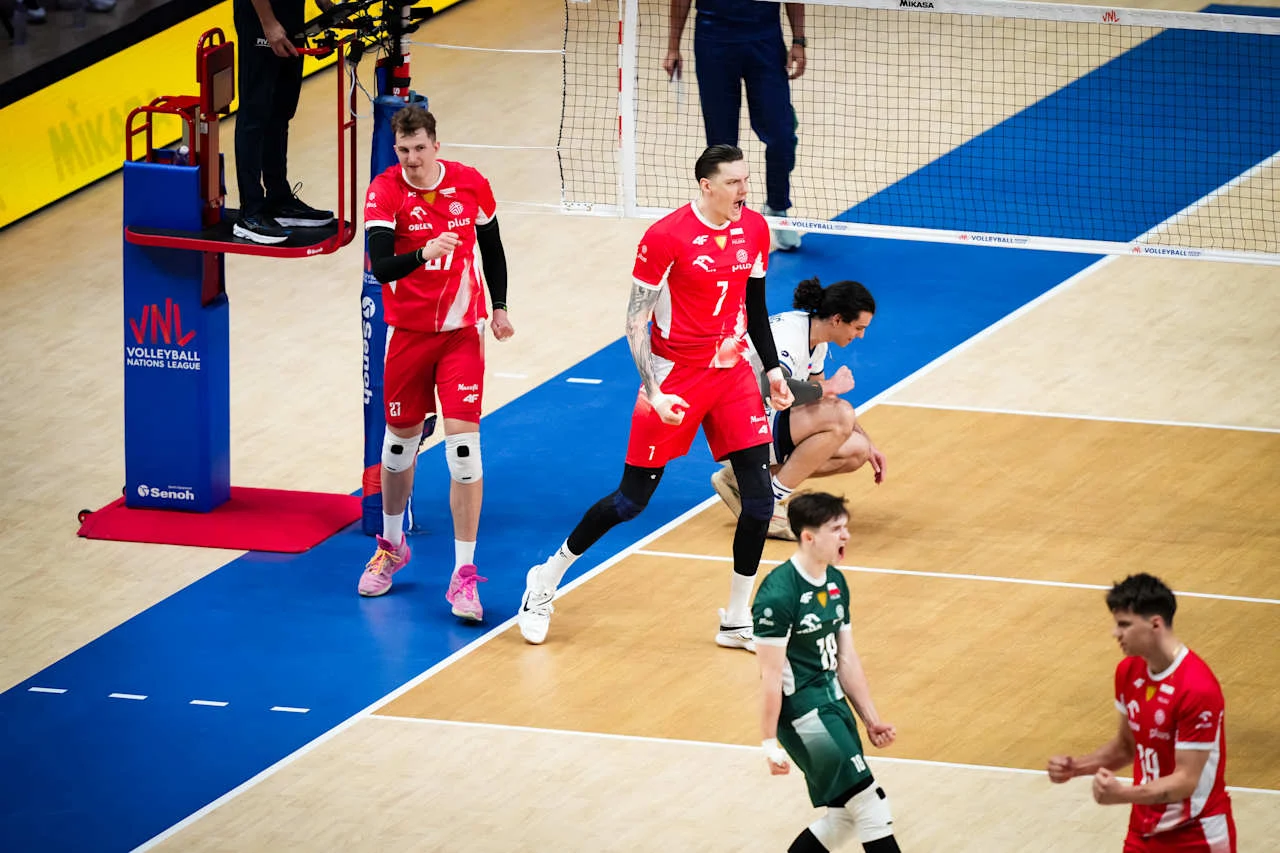 Dwóch siatkarzy w czerwonych strojach wyraża radość po zdobyciu punktu, podczas gdy zawodnicy przeciwnej drużyny wyglądają na rozczarowanych; sędzia obserwuje boisko z podestu, a całość rozgrywa się na profesjonalnej hali sportowej.
