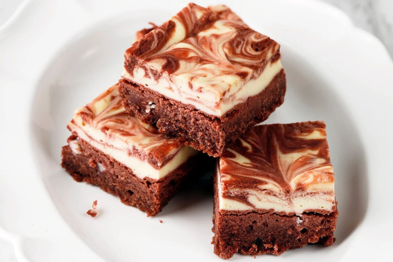 Brownie z polewą sernikową to eksplozja smaku Trzy kawałki brownie z marmurkową warstwą sernikową ułożone jeden na drugim na białym talerzu.