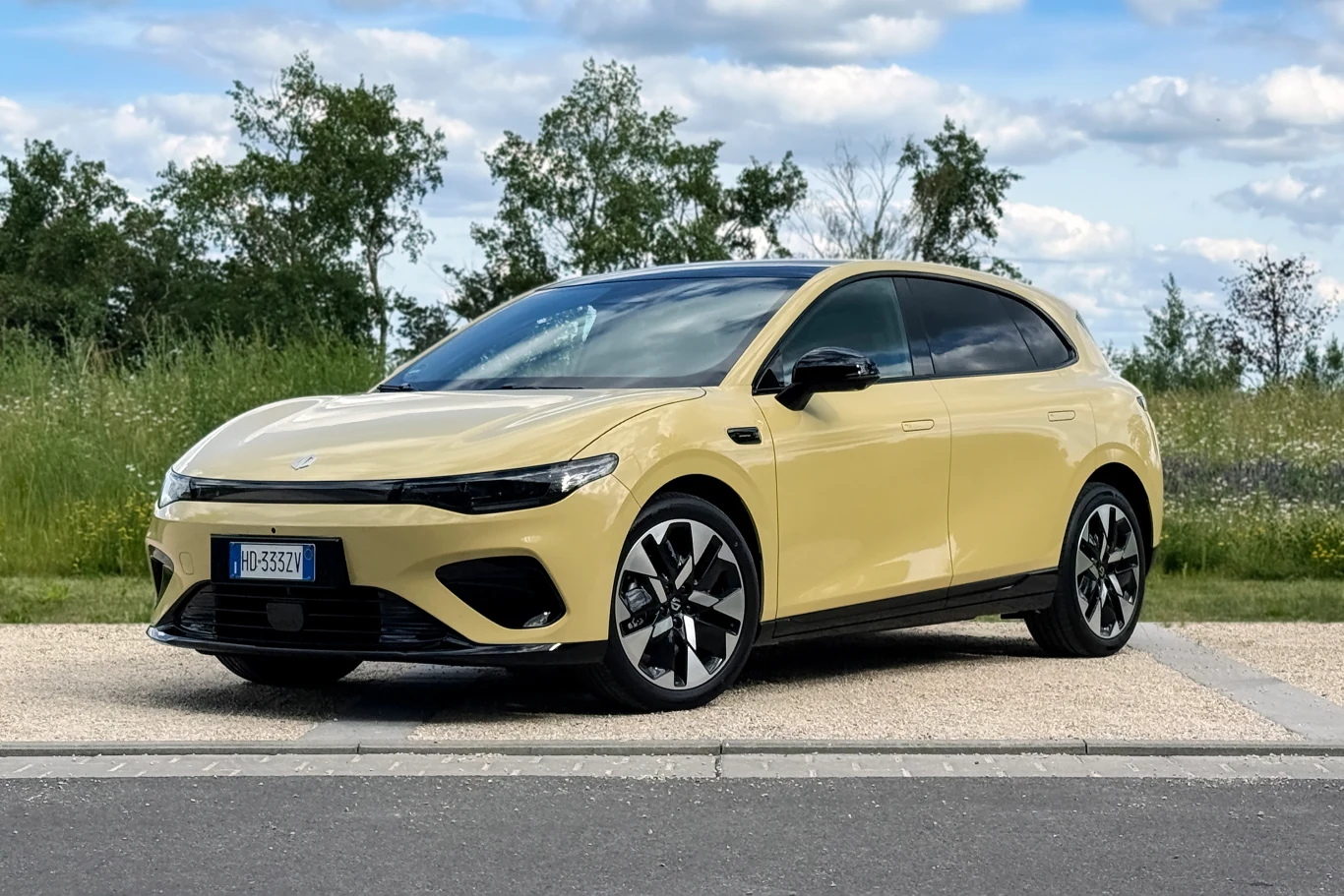 Jasnożółty nowoczesny samochód elektryczny z czarnym dachem i aerodynamicznymi felgami stoi na poboczu asfaltowej drogi, w tle roślinność i niebo z lekkim zachmurzeniem.