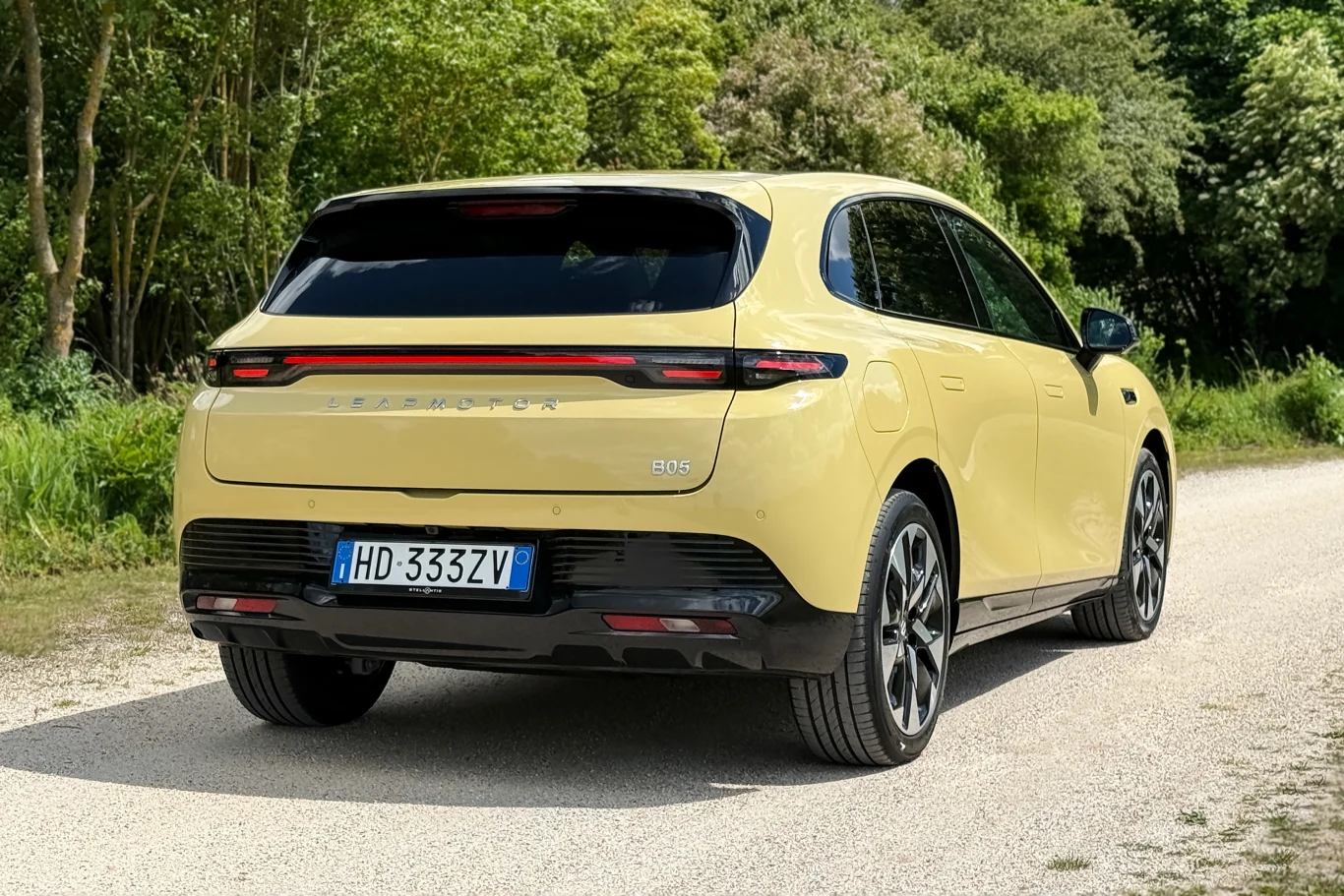 Jasnożółty nowoczesny samochód elektryczny o aerodynamicznym kształcie, czarnym dachu i przyciemnianych tylnych szybach, zaparkowany na szutrowej drodze otoczonej zielenią.