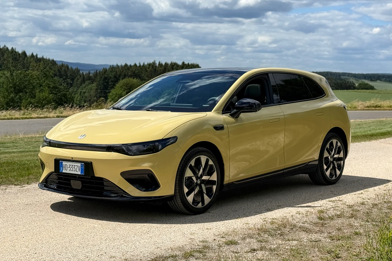 Jasnożółty samochód elektryczny o nowoczesnej sylwetce, ustawiony na poboczu asfaltowej drogi, otoczony zielenią i polami ze wzgórzami w tle.