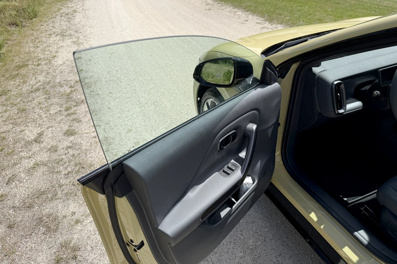 Otwarte przednie drzwi samochodu z szybą całkowicie opuszczoną w głąb drzwi, widoczna część wnętrza, w tym lusterko boczne i fragment fotela kierowcy, auto stoi na szutrowej drodze obok trawiastego pobocza.