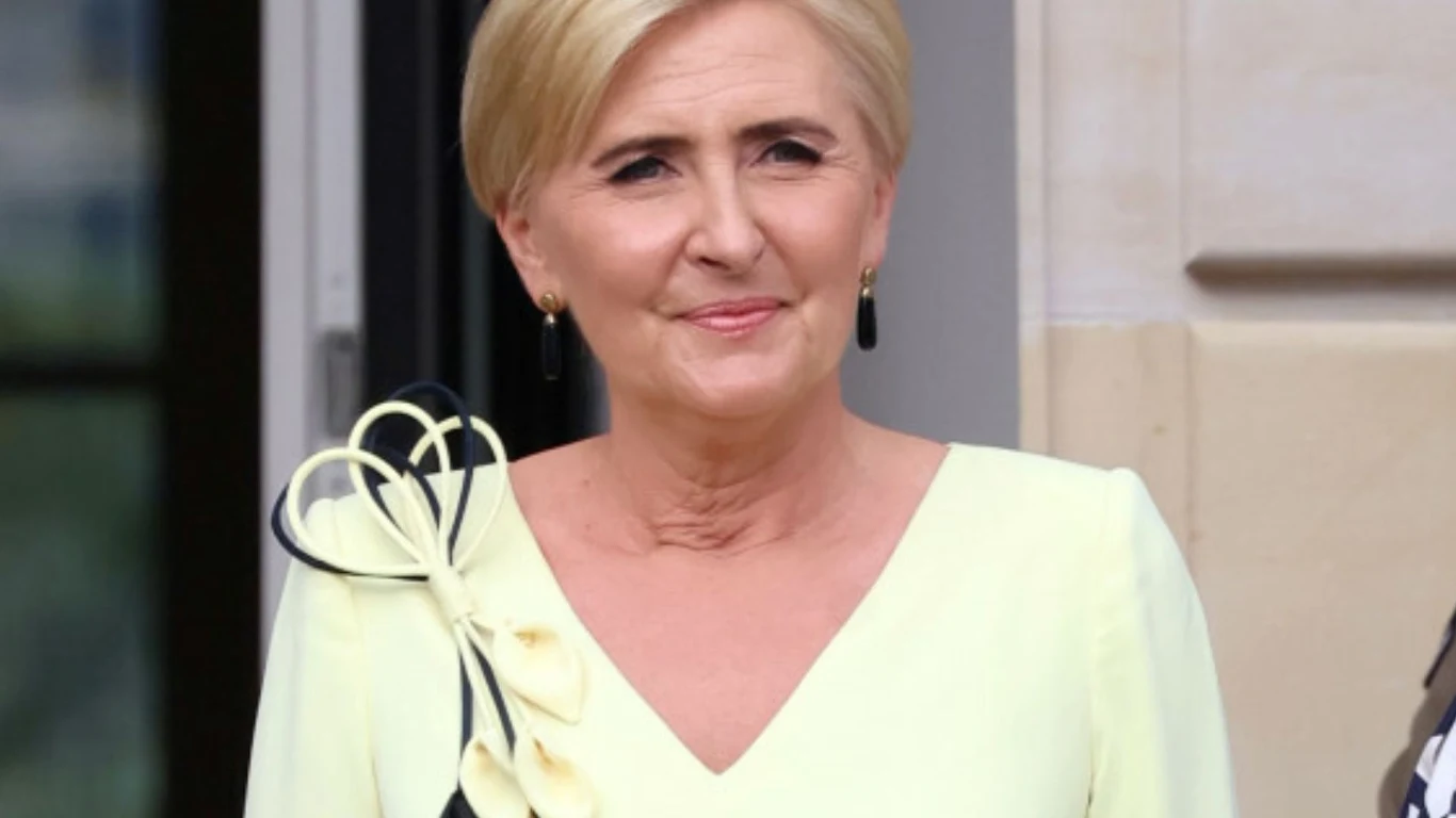 Agata Duda Agata Duda w jasnożółtej sukience z kokardą na ramieniu i krótkiej fryzurze, uśmiechnięta na tle jasnej ściany budynku.