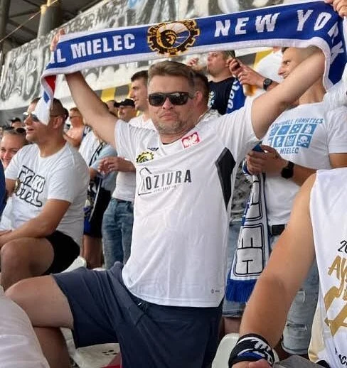 Mężczyzna w okularach przeciwsłonecznych, ubrany w biały t-shirt z logo klubu, trzyma rozwinięty szalik z napisem 'STAL MIELEC NEW YORK' na stadionie w otoczeniu kibiców.