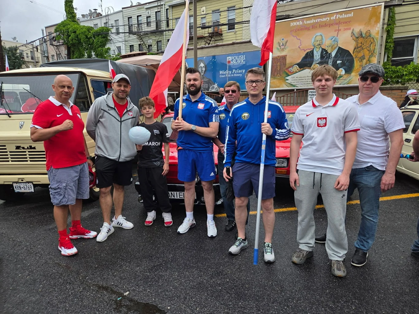 Grupa mężczyzn i chłopiec w sportowych ubraniach oraz koszulkach z polskim godłem stoi razem, trzymając dwie polskie flagi na tle zabytkowych samochodów; za nimi widoczny jest mural upamiętniający 100-lecie odzyskania niepodległości przez Polskę.