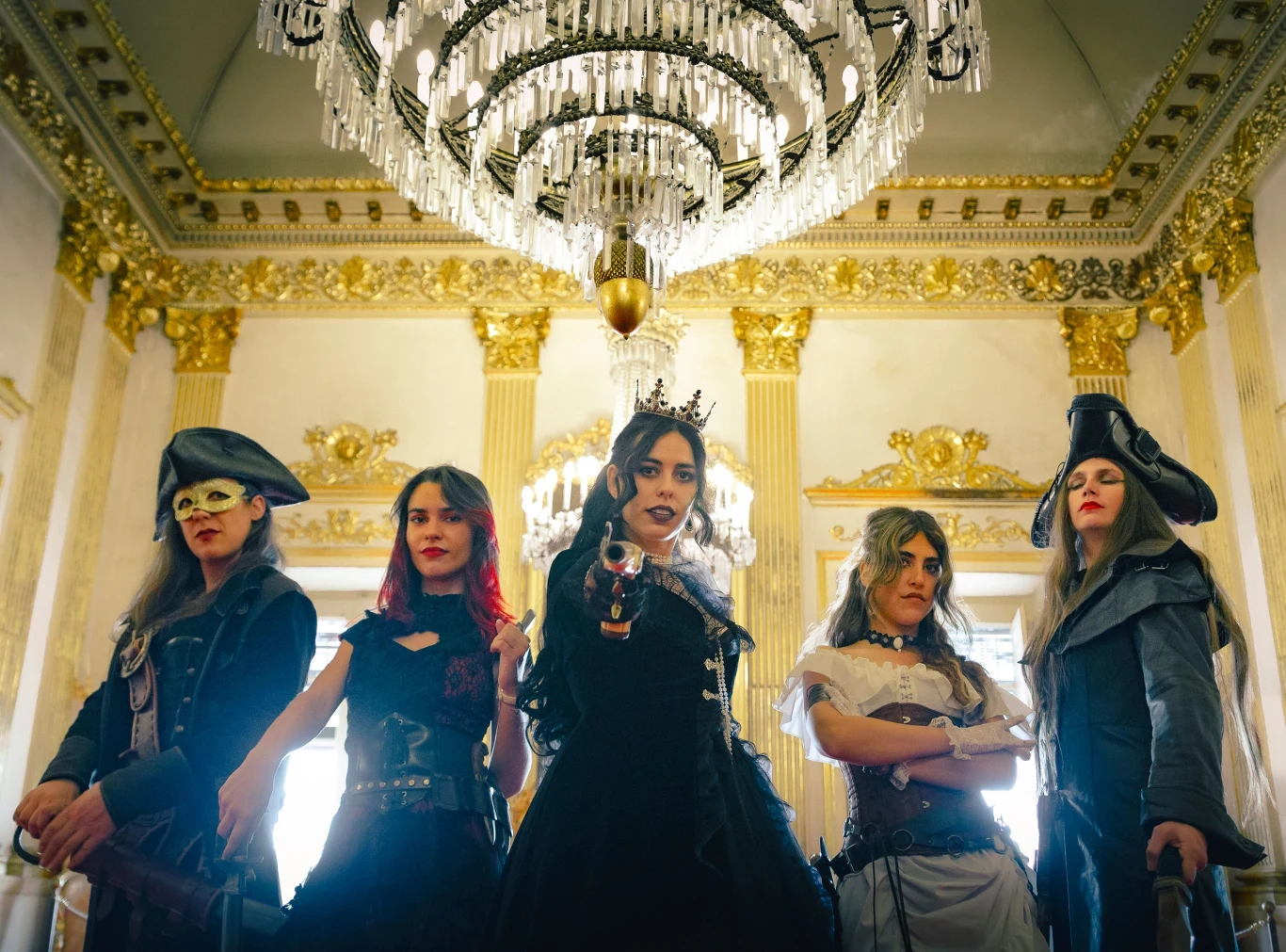 Pirate Queen w nowym składzie Pięć kobiet z Pirate Queen w steampunkowo-pirackich kostiumach pozuje pod żyrandolem w zdobionej sali.