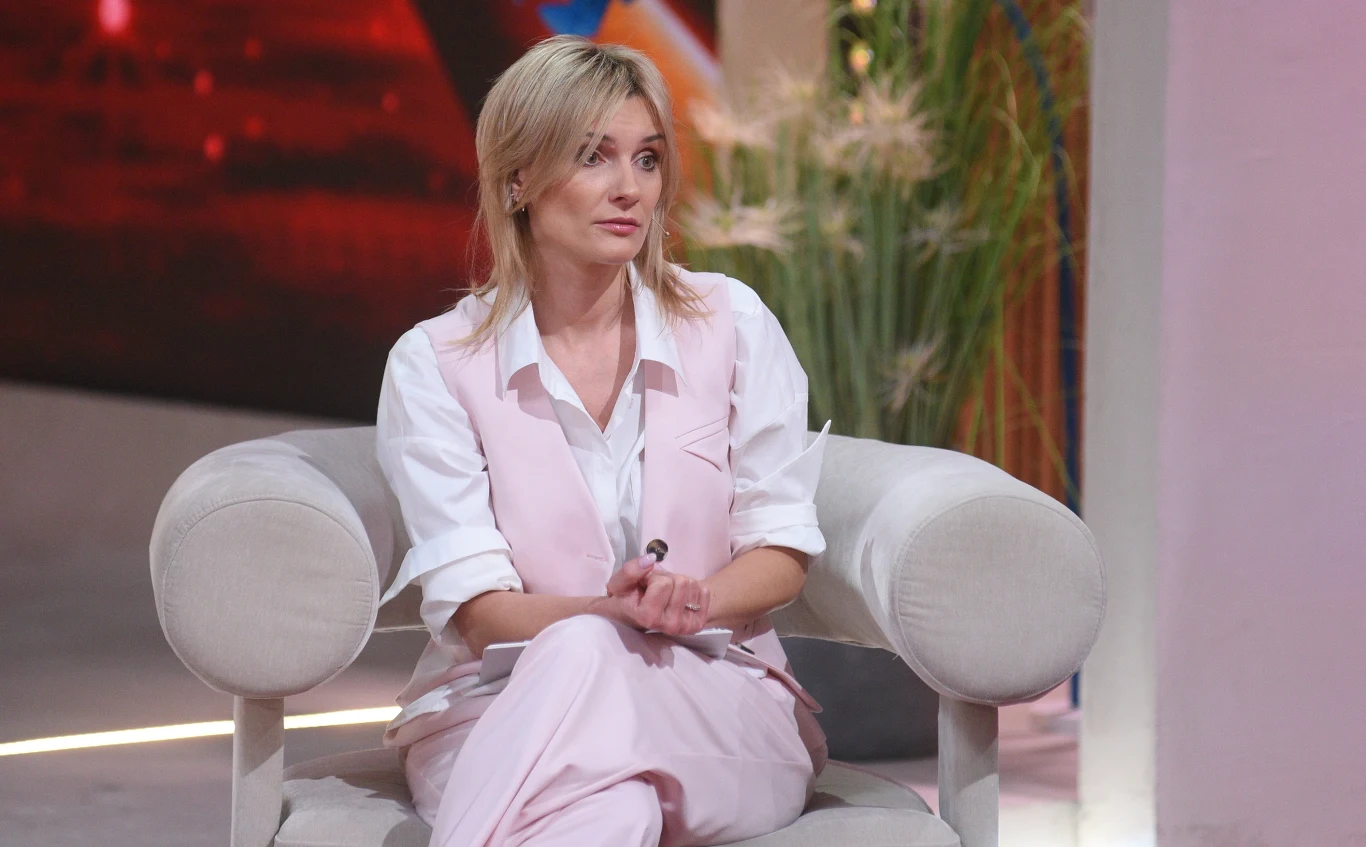 Anna Lewandowska Prezenterka Anna Lewandowska w jasnoróżowym garniturze, siedząca na fotelu w nowoczesnym studio telewizyjnym.