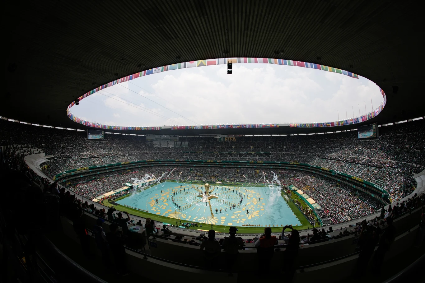 Pełny stadion z tłumem widzów, kolorową scenografią na murawie i uczestnikami widowiskowej ceremonii na środku boiska.