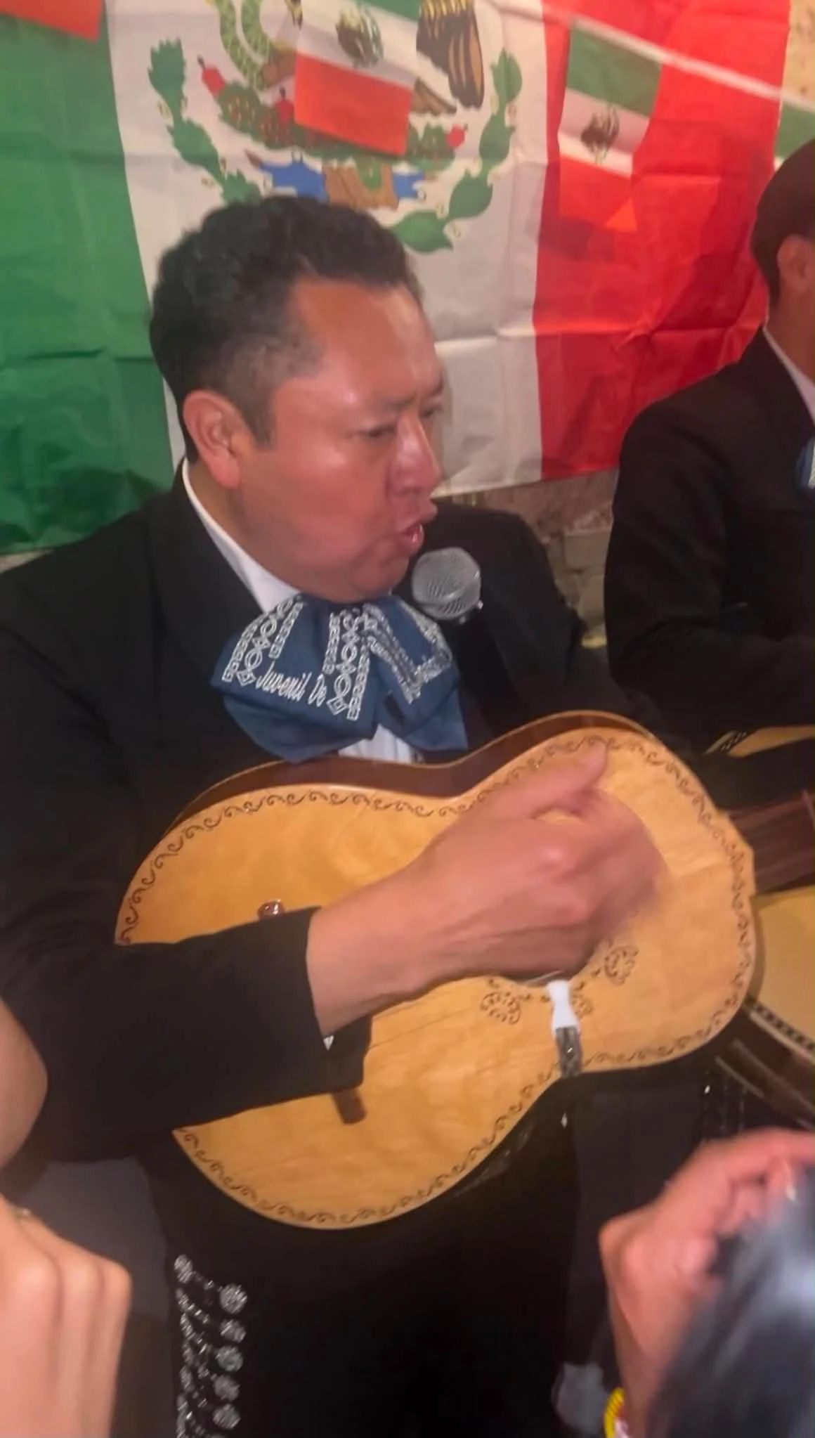 Mężczyzna ubrany w tradycyjny strój mariachi gra na gitarze i śpiewa do mikrofonu, w tle widoczna duża flaga Meksyku.