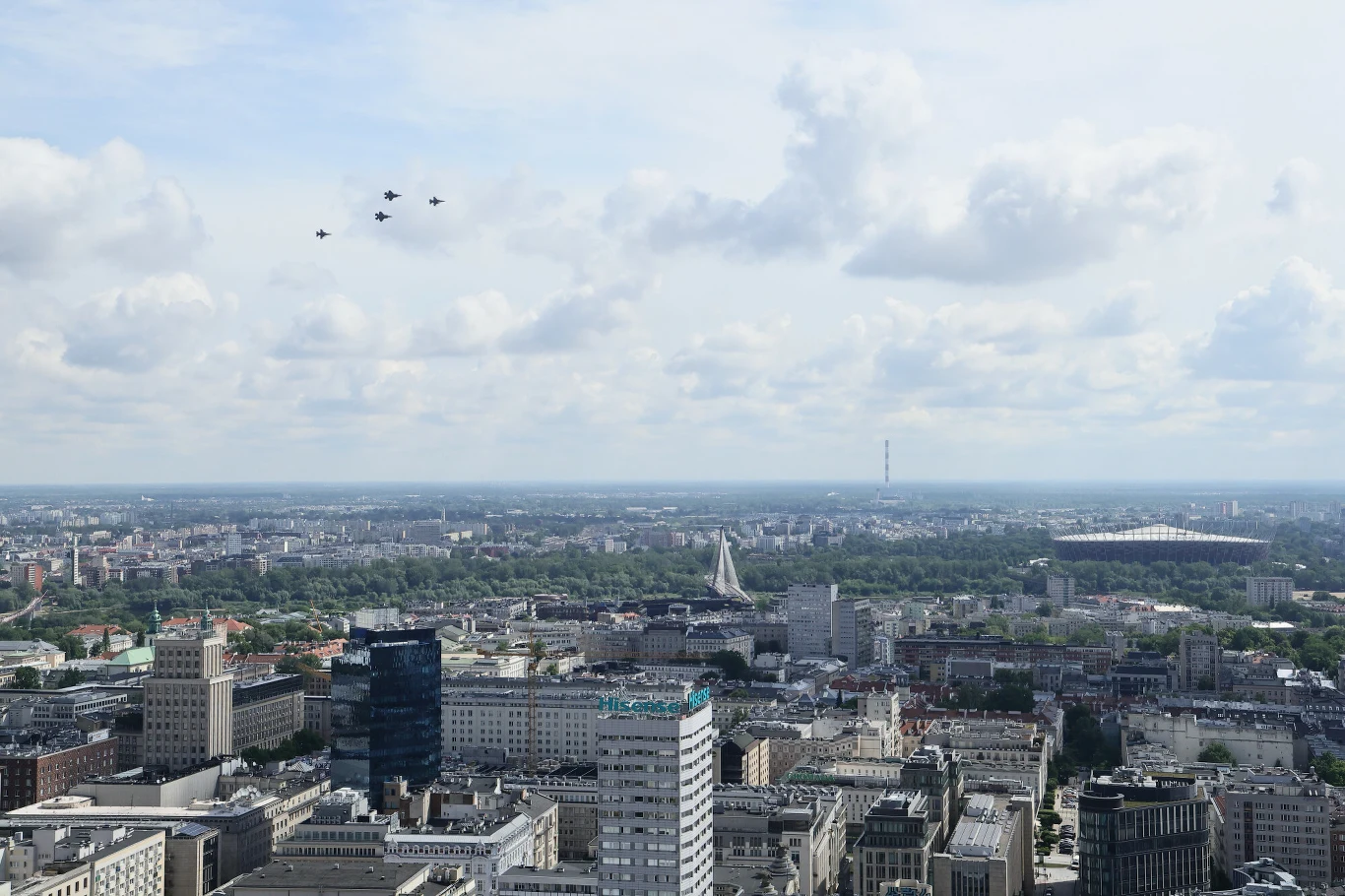 Panorama Warszawy z widocznymi nowoczesnymi i starszymi budynkami, czterema myśliwcami lecącymi w szyku na niebie oraz Stadionem Narodowym w tle.