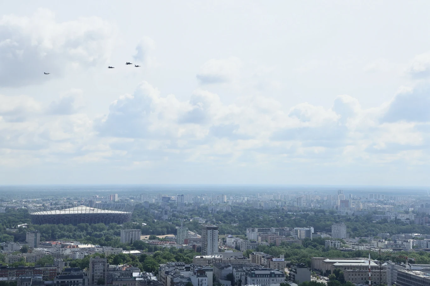 Widok na panoramę miasta z dużym stadionem po lewej stronie i formacją czterech samolotów lecących nad gęsto zabudowaną dzielnicą.