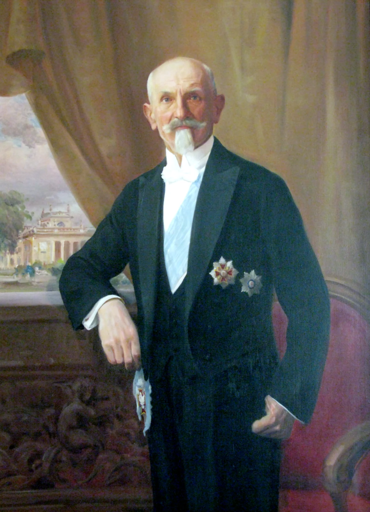 Starszy mężczyzna w eleganckim fraku z orderami przypiętymi do marynarki, stojący przy ozdobionym stole na tle grubych zasłon i widoku na okazały pałac za oknem.
