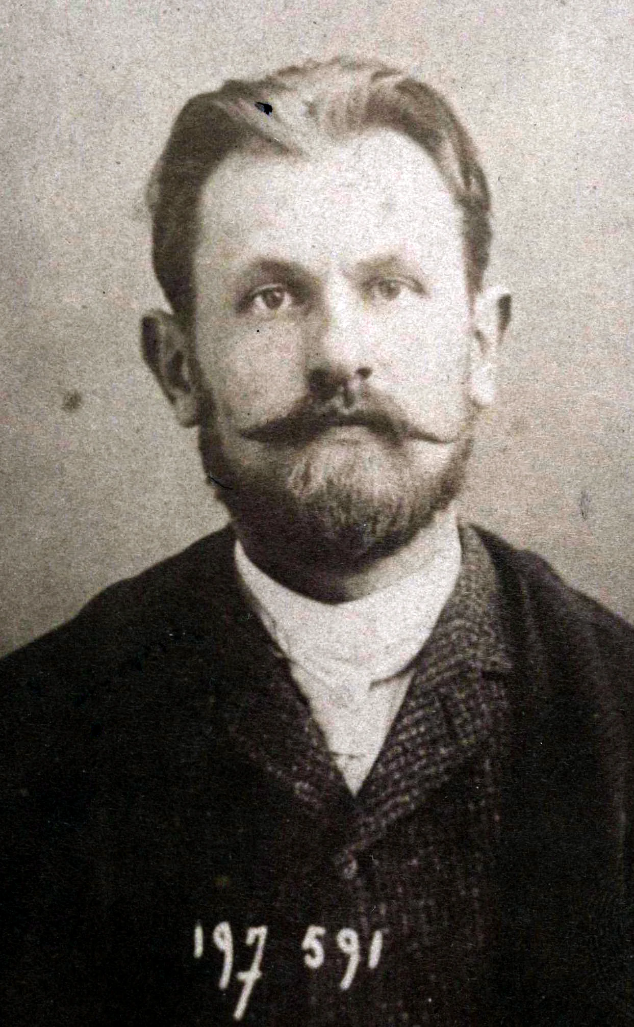 Portret mężczyzny z krótkimi włosami oraz charakterystycznym, gęstym wąsem i brodą, ubrany w garnitur z białą koszulą, na dole widoczny numer identyfikacyjny.