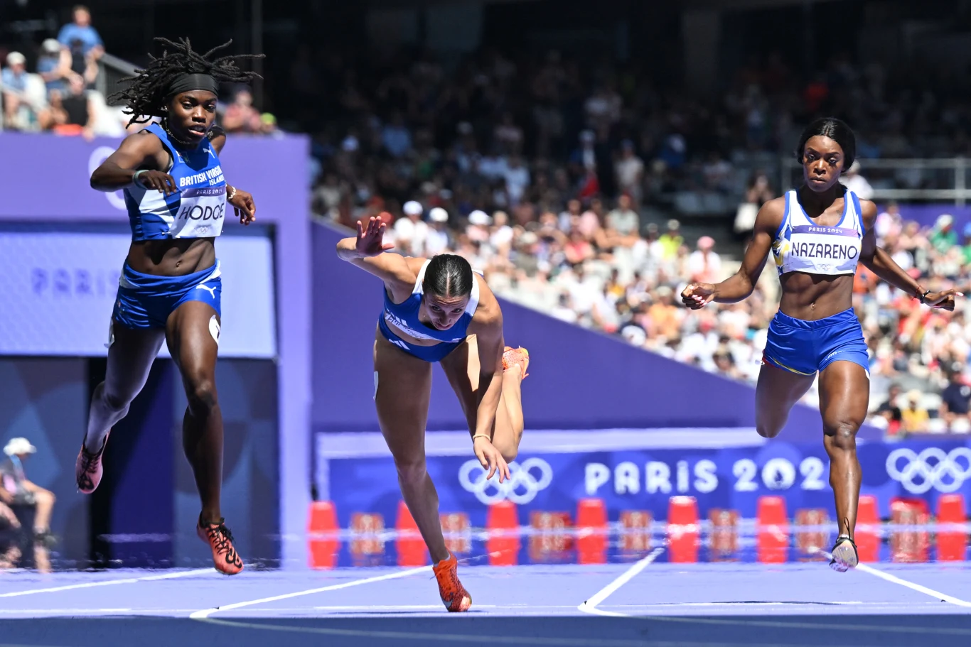 Trzy zawodniczki biegnące po bieżni, jedna z nich wykonuje dynamiczny rzut na linię mety, a w tle widoczny napis Paris 2024 i logo igrzysk olimpijskich.