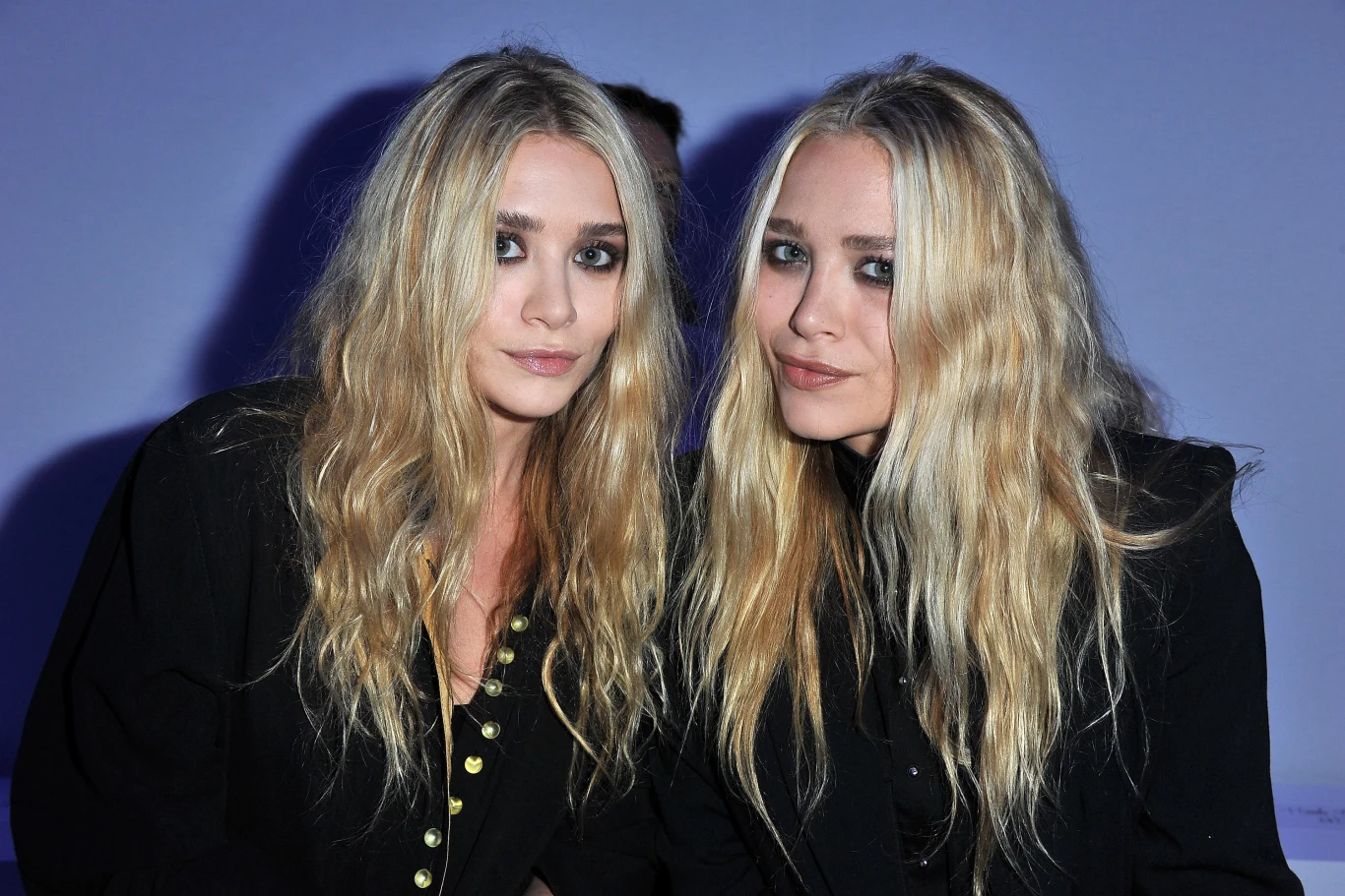 Mary-Kate i Ashley Olsen Dwie kobiety o długich, blond włosach i podobnych rysach twarzy siedzą obok siebie, ubrane w ciemne marynarki, spoglądając bezpośrednio w obiektyw na jasnym tle.