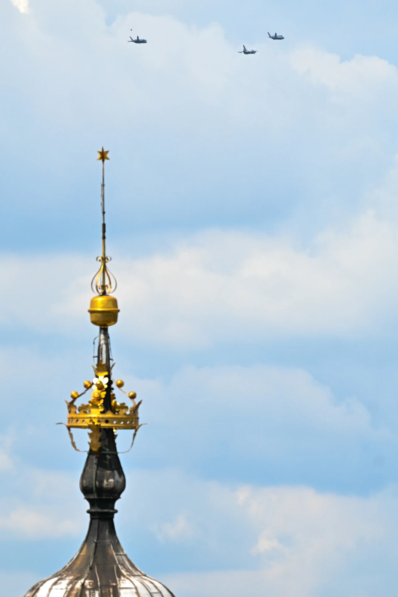 Wieża zwieńczona złotą koroną z wysokim ornamentem oraz trzy samoloty lecące w formacji na tle błękitnego, lekko zachmurzonego nieba.