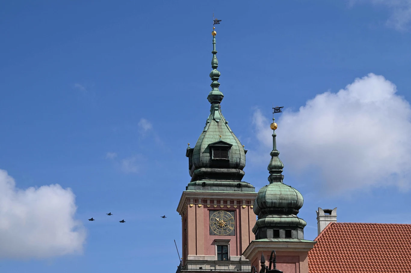 Zabytkowa wieża z zegarem i zielonymi kopułami kontrastuje z niebieskim niebem, w tle widoczny przelot czterech myśliwców.