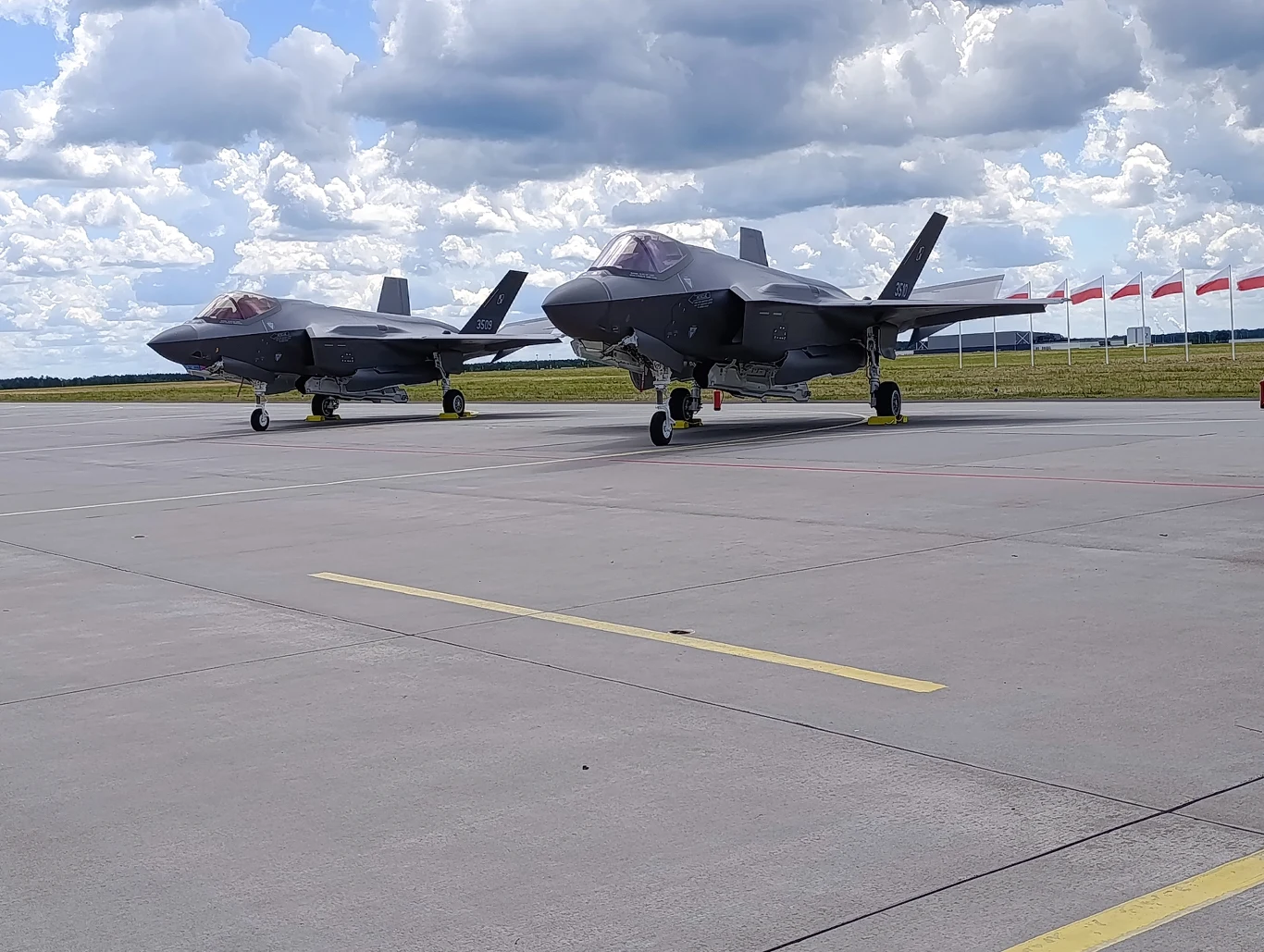 Dwa nowoczesne myśliwce F-35 ustawione na pasie lotniska, na tle nieba z gęstymi chmurami i rzędu biało-czerwonych flag po prawej stronie.