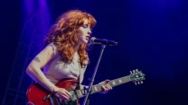 Kobieta o rudych włosach grająca na czerwonej gitarze elektrycznej i śpiewająca do mikrofonu na scenie koncertowej oświetlonej niebieskim światłem.