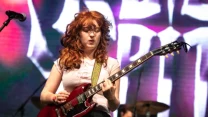 Młoda kobieta z rudymi, kręconymi włosami gra na czerwonej gitarze elektrycznej na scenie koncertowej, w tle perkusistka i barwne oświetlenie estradowe.