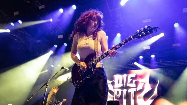 Gitarzystka występująca na scenie podczas koncertu, dynamiczne światła i logo zespołu Die Spitz w tle.