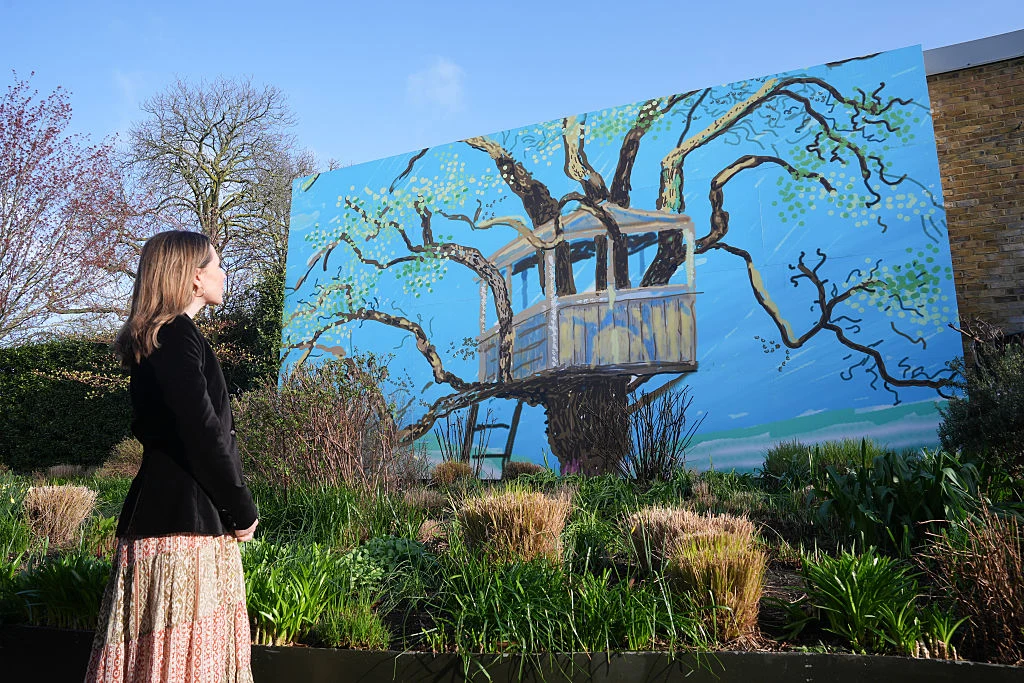 Kobieta w długiej, wzorzystej spódnicy patrząca na dużą mural przedstawiający domek na drzewie, otoczona zielenią i krzewami.