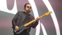 Mężczyzna w ciemnych okularach i czarnej skórzanej kurtce gra na gitarze basowej na tle dużej sceny oświetlonej światłem dziennym.
