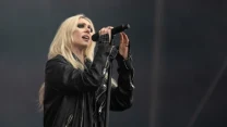 Kobieta z długimi blond włosami w czarnej skórzanej kurtce śpiewa do mikrofonu na scenie podczas występu muzycznego.