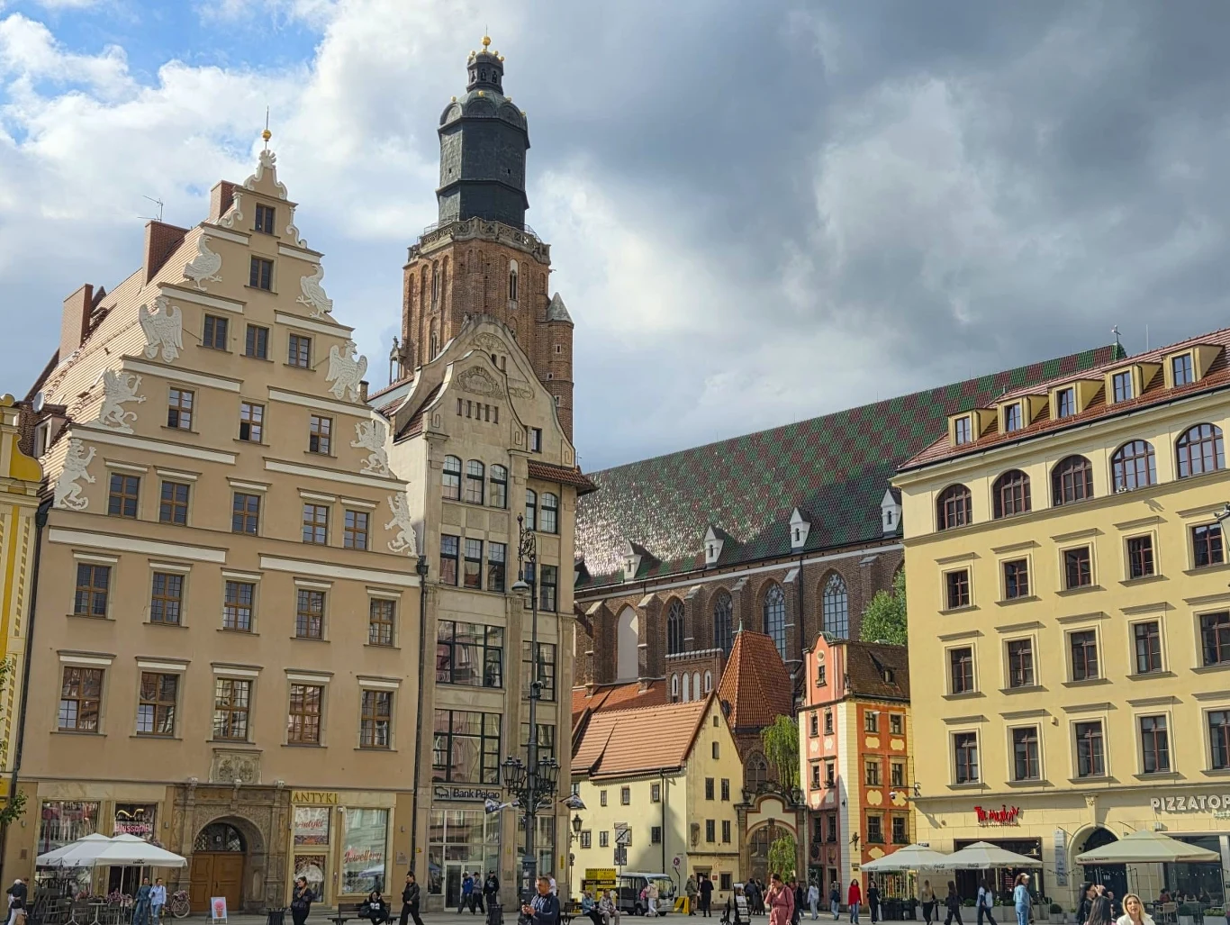 Kolorowe kamienice otaczające plac, w tle widoczny gotycki kościół z wysoką wieżą oraz charakterystycznym, dachówkowym dachem, poniżej spacerujący ludzie i ogródki kawiarniane.