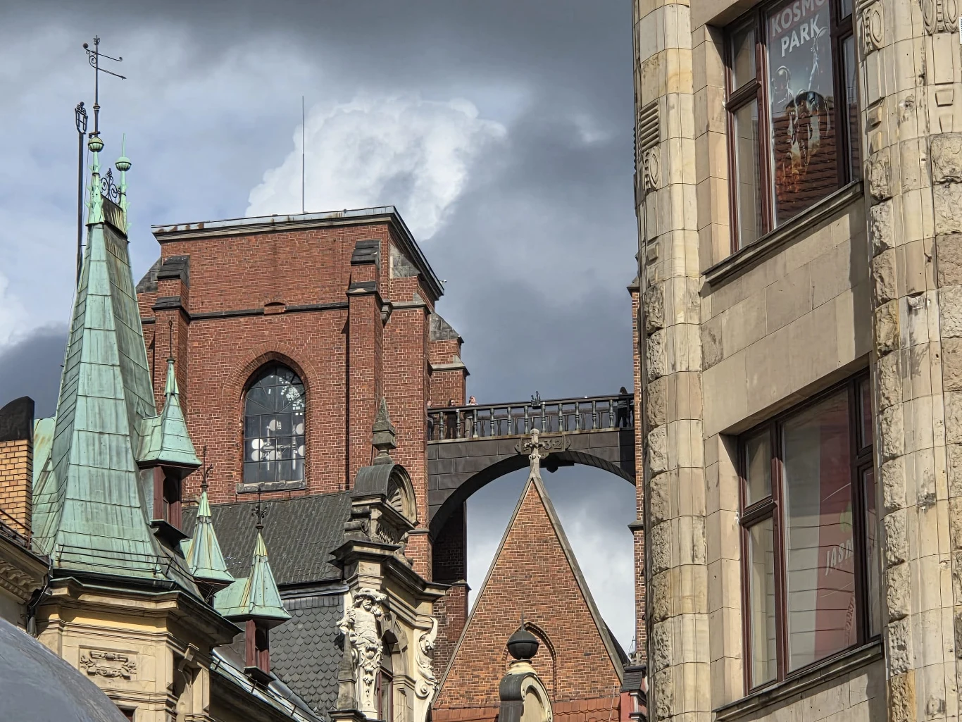 Ceglana wieża kościelna połączona wysoką arkadą z sąsiednim budynkiem, w otoczeniu zabytkowych kamienic z zielonymi i szarymi dachami, pod dramatycznym, pochmurnym niebem.