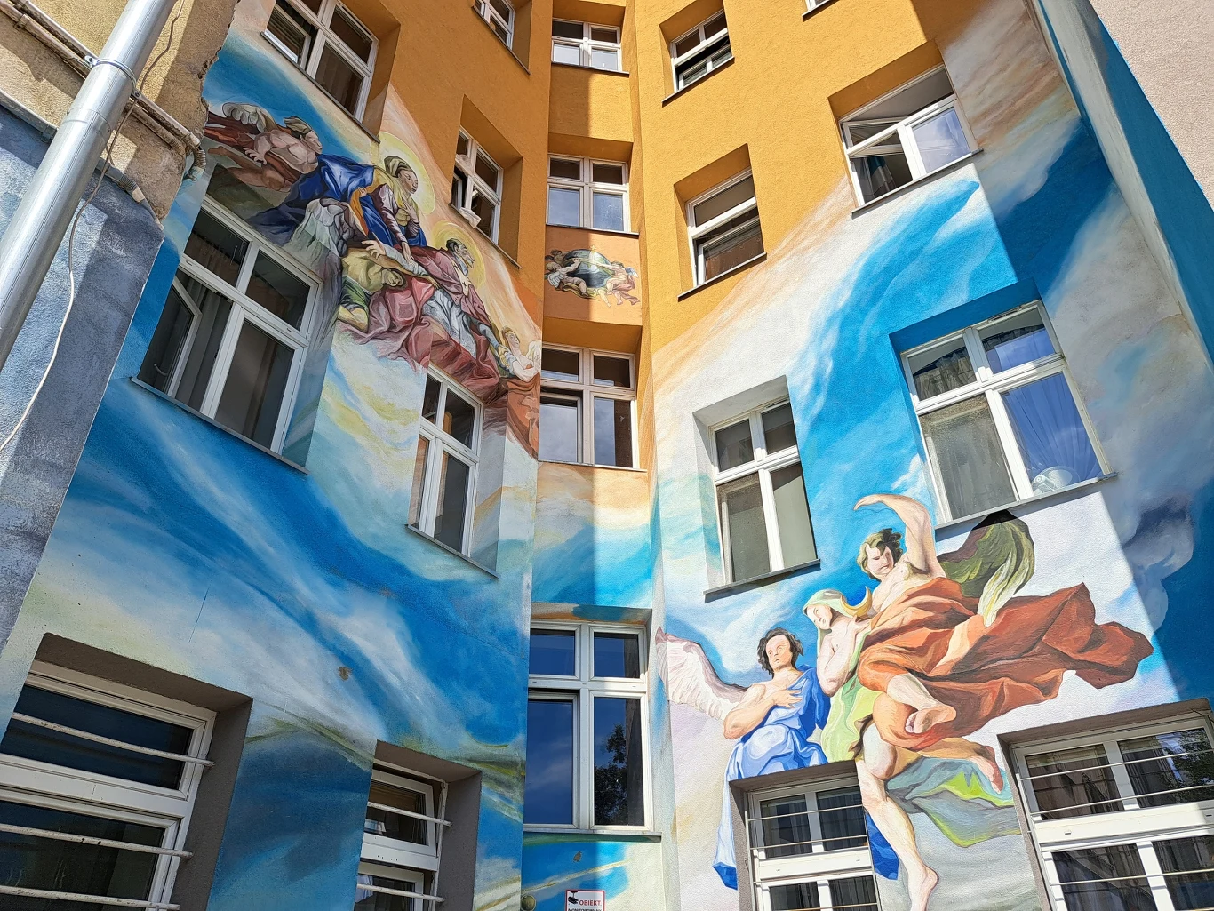 Kolorowy mural na ścianie budynku przedstawiający postacie ubrane w antyczne szaty, unoszące się wśród błękitnych obłoków, z detalami nawiązującymi do sztuki renesansowej.