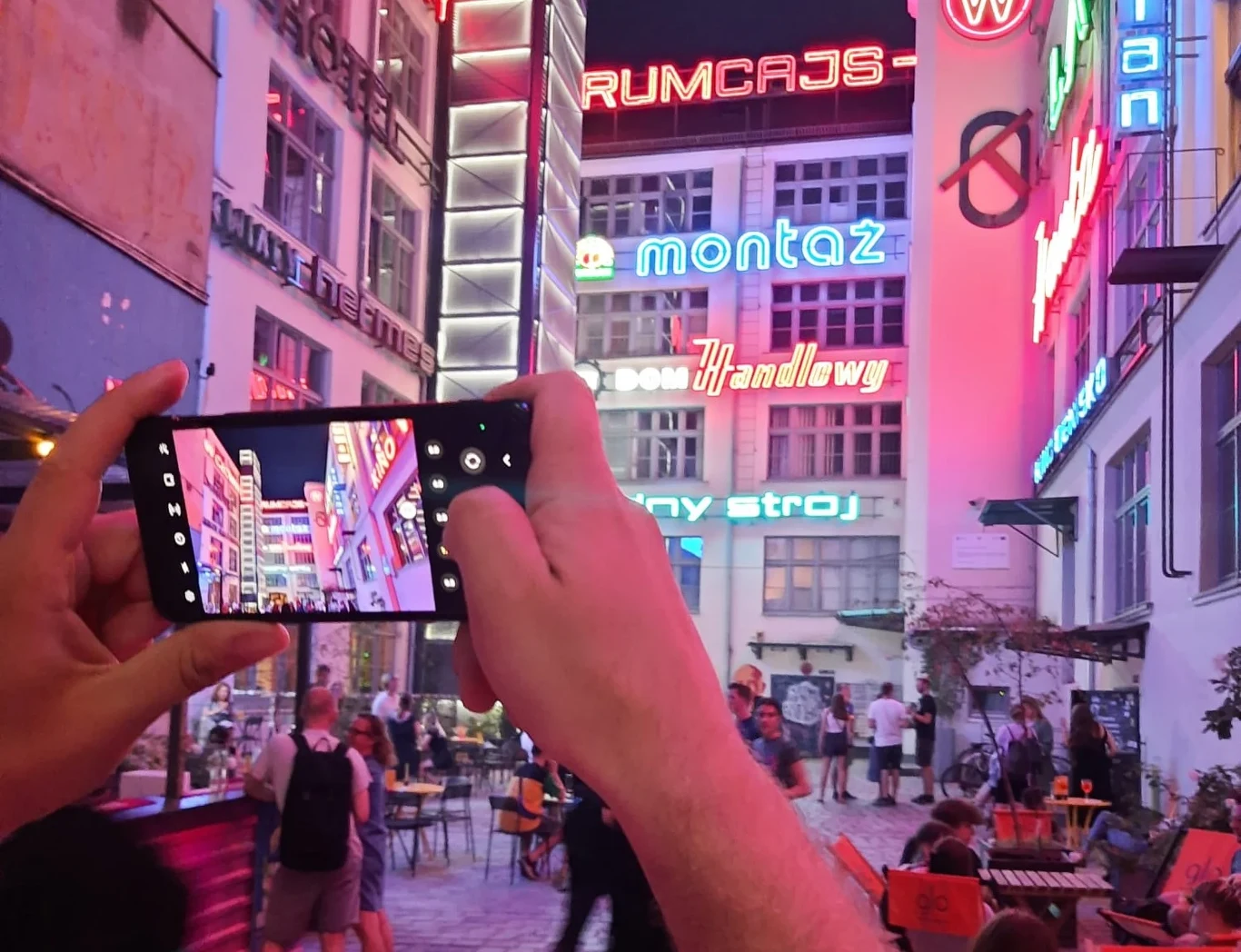 Ręce trzymające smartfon, którym robione jest zdjęcie kolorowych neonów na ścianach w miejskiej przestrzeni z licznymi napisami i ludźmi siedzącymi przy stolikach kawiarni na zewnątrz.