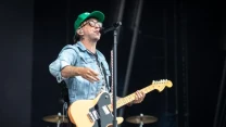 Muzyk w okularach i zielonej czapce z daszkiem gra na gitarze elektrycznej i śpiewa do mikrofonu na scenie koncertowej.