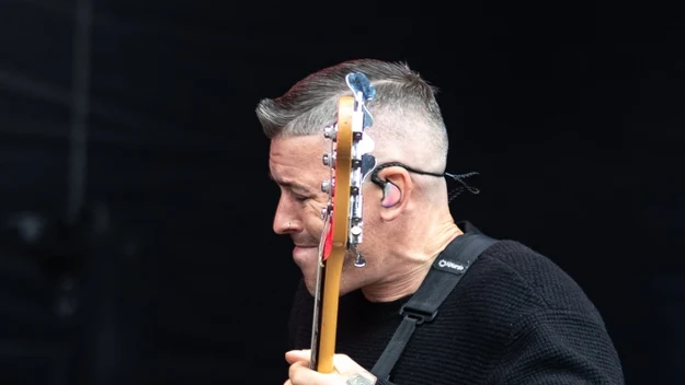 Mężczyzna z krótkimi, siwymi włosami gra na różowej gitarze basowej, ubrany w czarny sweter i czarne spodnie, ma założony mikrofon nagłowny.