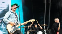 Trzech muzyków podczas energetycznego występu na scenie; wokalista z gitarą elektryczną w okularach i czapce z daszkiem, perkusista grający dynamicznie na bębnach oraz basista zwrócony bokiem do widowni.