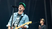 Mężczyzna w zielonej czapce oraz okularach śpiewa i gra na gitarze elektrycznej na scenie podczas koncertu, w tle drugi muzyk również z gitarą.