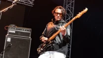 Mężczyzna w okularach przeciwsłonecznych grający na gitarze basowej na scenie koncertowej, obok wzmacniacza i sprzętu nagłośnieniowego.