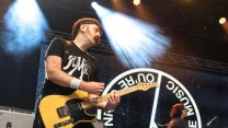 Gitarzysta z żółtą gitarą elektryczną występuje na scenie obok klawiszowca, w tle duże okrągłe logo zespołu oraz światła sceniczne.