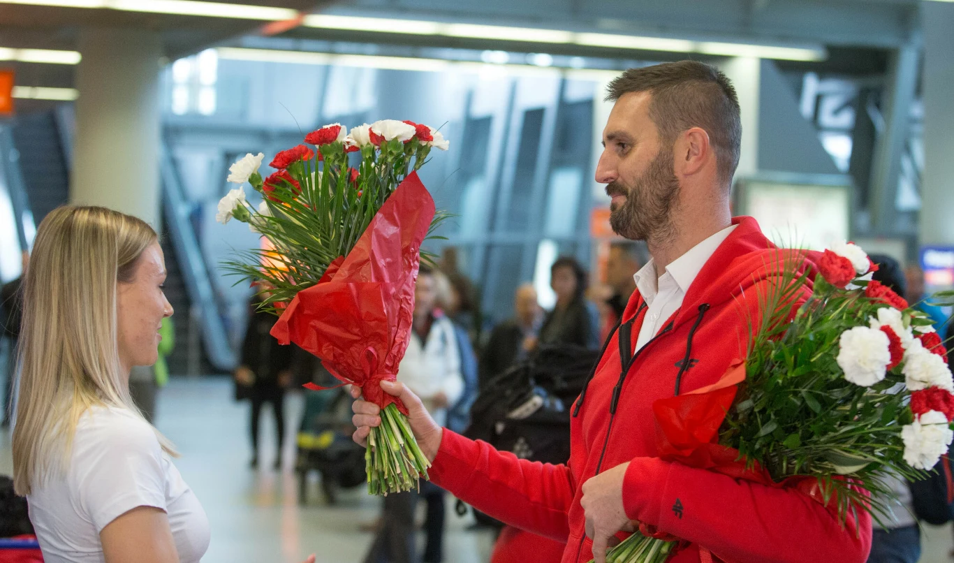 Mężczyzna ubrany w czerwoną bluzę wręcza bukiet czerwonych i białych goździków kobiecie, oboje są uśmiechnięci, znajdują się w hali lotniska.