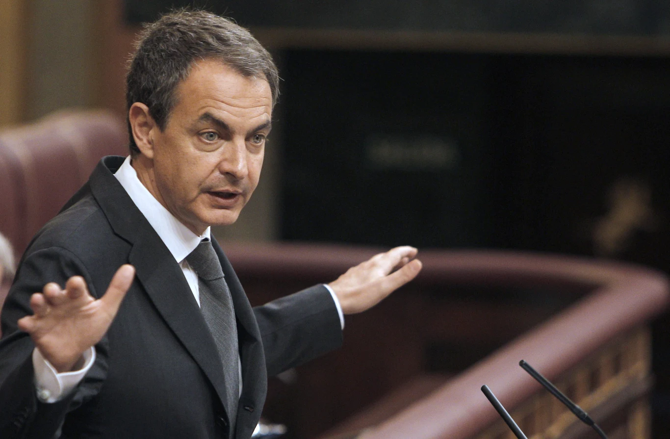 Jose Luis Rodriguez Zapatero w garniturze przemawia przy mównicy, gestykulując rękoma do słuchaczy.