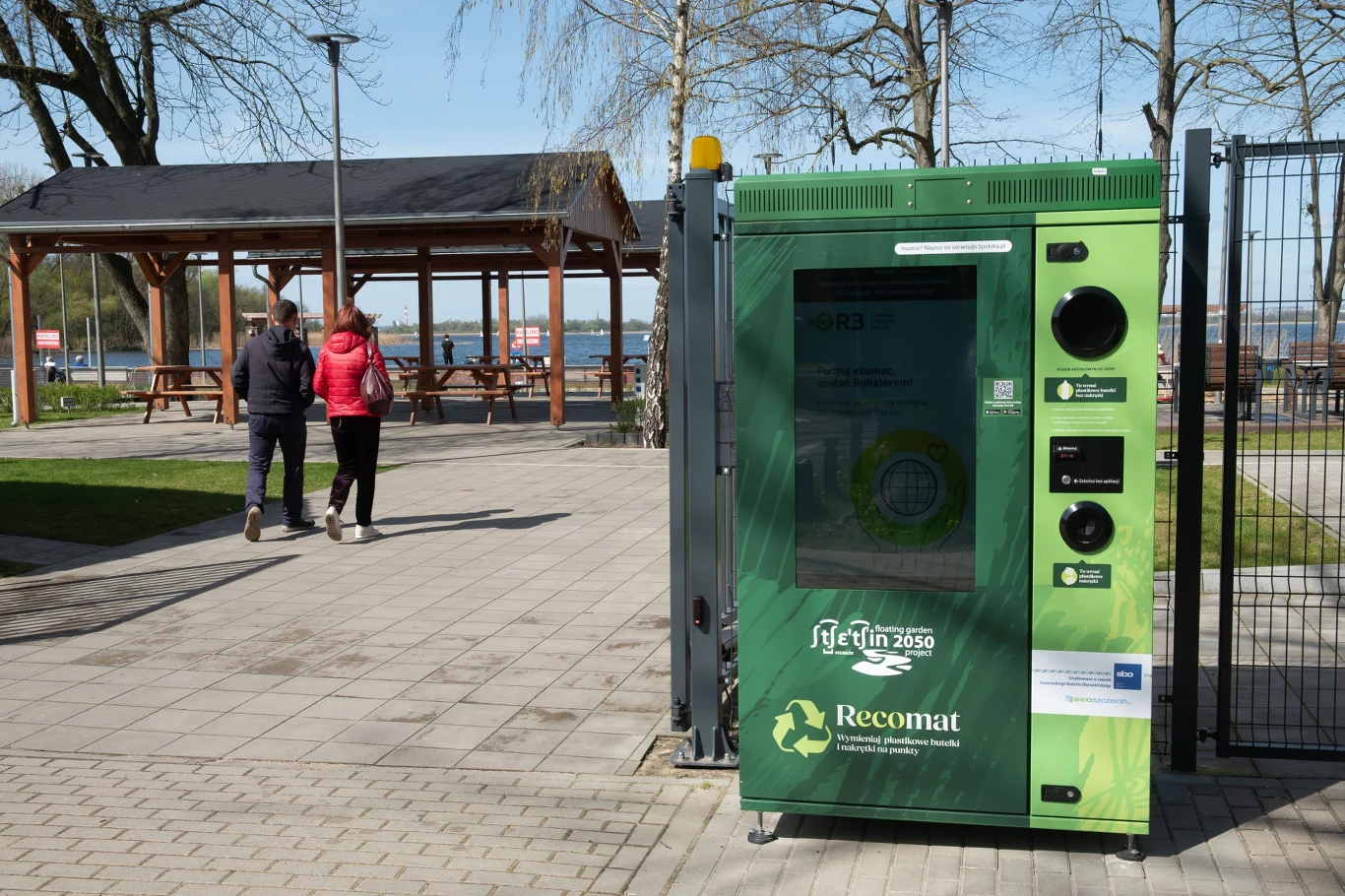 Recyklomat w Szczecinie ustawiony przy ogrodzeniu, w tle dwie osoby i altana z ławkami.