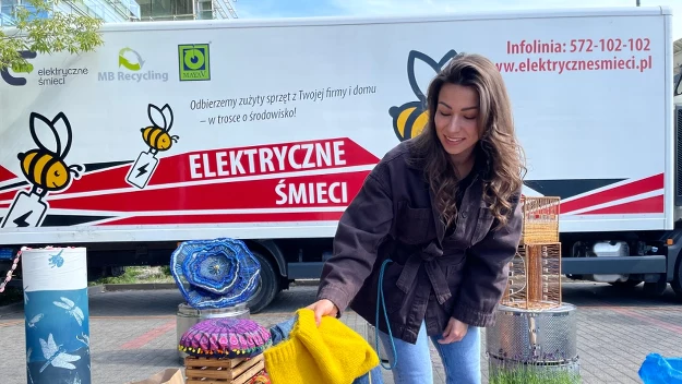 Kobieta wybierająca ubrania przy stoisku z kolorowymi pudełkami i torebkami, w tle samochód firmy zajmującej się odbiorem elektroodpadów ozdobiony charakterystyczną grafiką pszczoły.