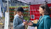 Dwie kobiety podczas rozmowy na stoisku z roślinami, jedna z nich wręcza drugiej sadzonki lawendy, w tle ustawione rzędy doniczek i baner promocyjny.