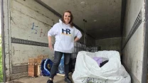 Kobieta w białej bluzie z napisem HR stoi w przestronnym wnętrzu ciężarówki obok dużego białego worka i trzech doniczek z lawendą; w tle widoczne są skrzynki i zwinięte dywany.