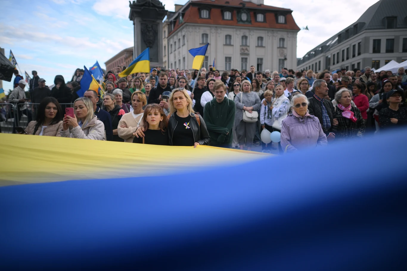 Tłum ludzi na placu z dużą flagą Ukrainy na pierwszym planie; widoczne także inne ukraińskie flagi, budynki w tle.