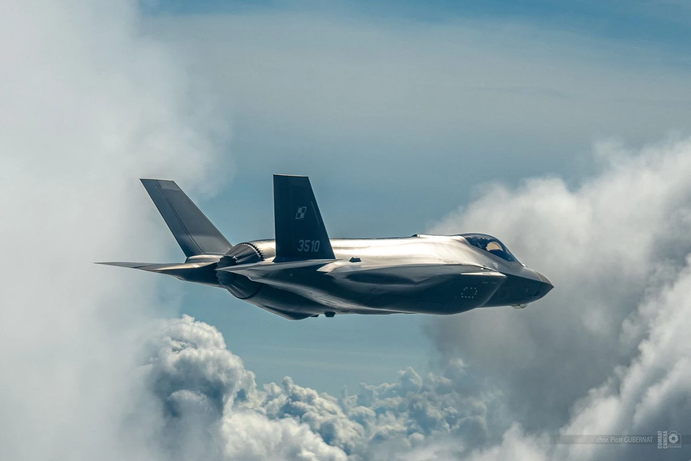 Nowoczesny myśliwiec wojskowy F-35 szybujący na wysokości, otoczony chmurami, widoczny z boku na tle jasnego nieba.