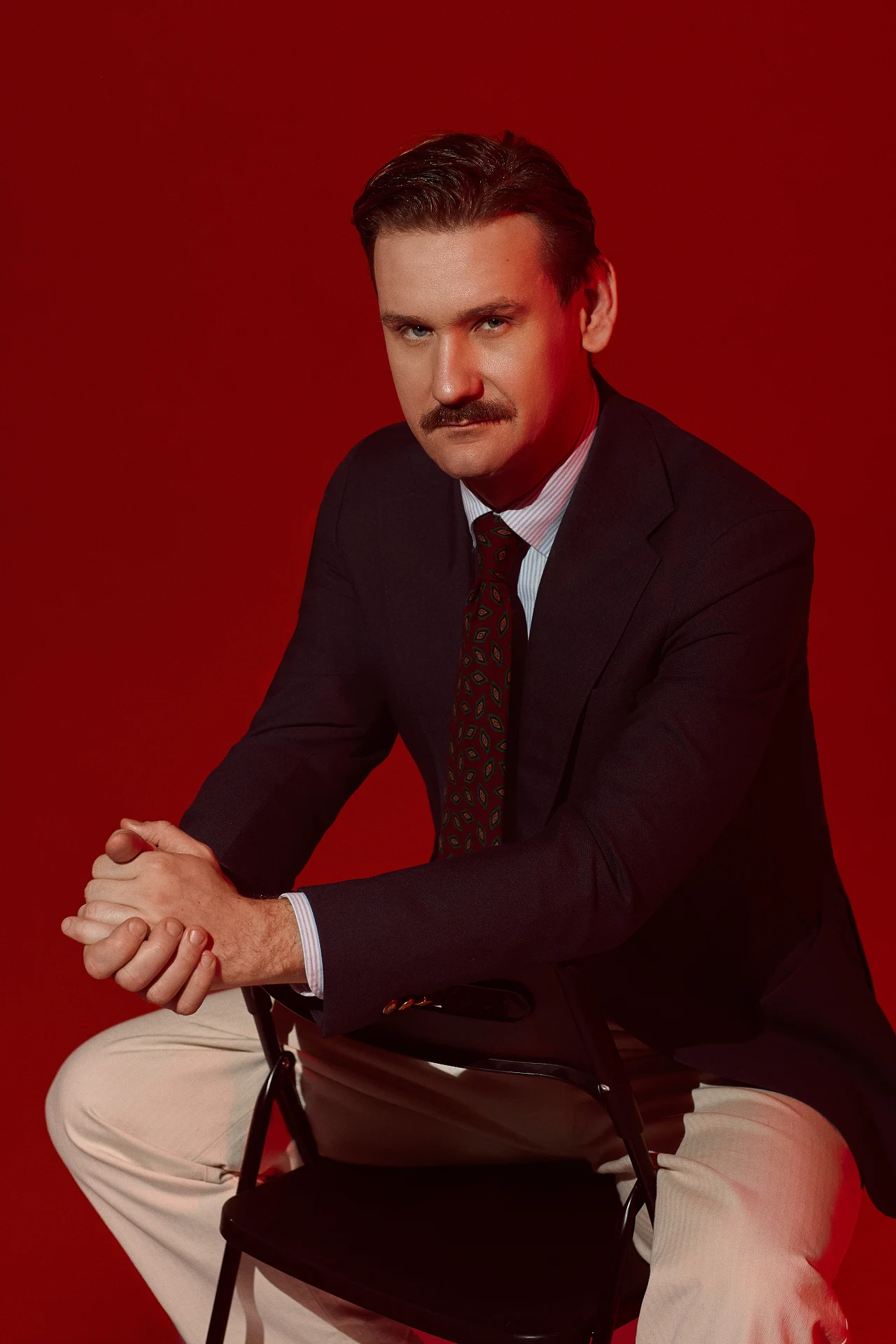 Jakub Dymek Mężczyzna z ciemnymi włosami i wąsem w eleganckiej marynarce, koszuli i krawacie, siedzi na czarnym krześle na tle jednolicie czerwonej scenografii.