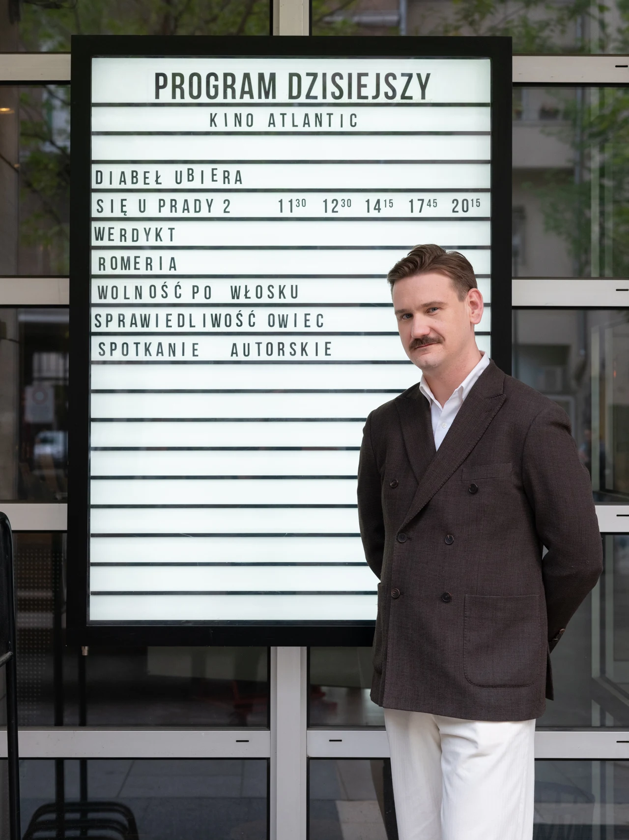Jakub Dymek Mężczyzna w eleganckim garniturze stoi przed planszą z repertuarem kina Atlantic, na której widoczne są tytuły filmów oraz godziny seansów.