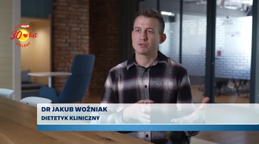 Mężczyzna w kraciastej koszuli siedzi przy stole i gestykuluje podczas wypowiedzi, obok wyświetlone są napisy informujące o jego imieniu, nazwisku i specjalizacji.