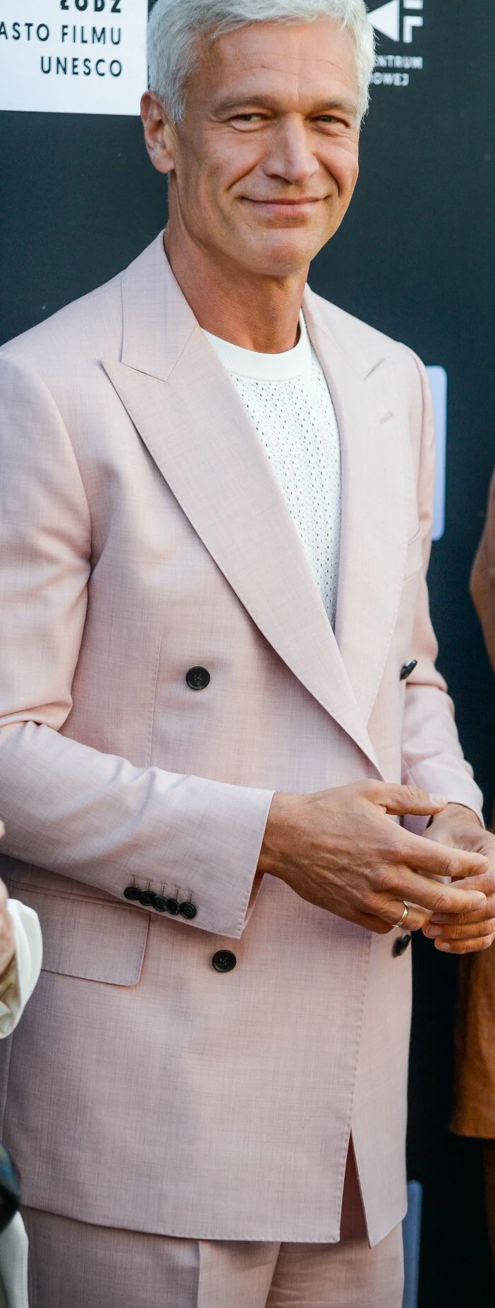 Michał Żebrowski w Łódzkiej Alei Gwiazd Elegancko ubrany mężczyzna w jasnoróżowym garniturze i białym swetrze stoi na tle czarnej ścianki z białymi napisami.